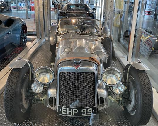 A shiny vintage car on display, featuring a chrome body, a distinctive front grille, and round headlights. The car is positioned within a glass enclosure, showcasing its design and detailing against a modern backdrop with additional vehicles visible in the background.