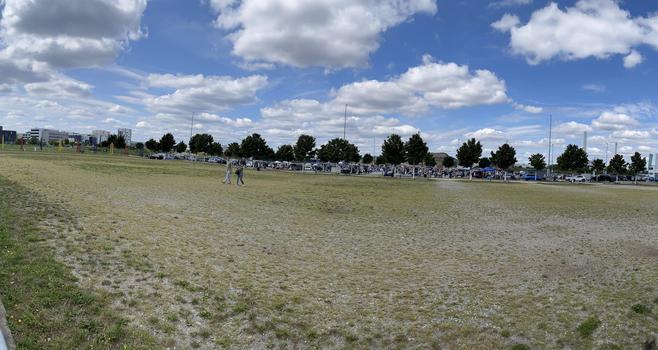 A panoramic view of an open field with a cloudy sky overhead. In the background, a crowd can be seen along a fence, with vehicles parked nearby. There are trees lining the area and colored poles set up in the field.