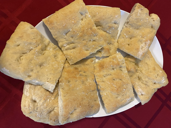 Seen from above. A white oval platter on a red tablecloth. Eight pieces of plain focaccia are arranged on the platter. One can see the dimples in the golden-brown bread and the Maldon salt flakes sprinkled on top.