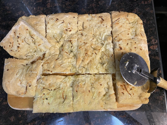 Seen from above. A wooden cutting board on a dark granite counter that is reflecting a lot of glare. On the board are twelve freshly cut golden-brown pieces of focaccia. A couple of pieces are angled for visual interest. Onion, rosemary, and Maldon salt flakes top the focaccia. To the lower right is a pizza cutter, resting against a couple of pieces.