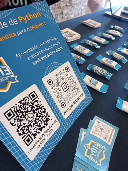 Visão mais próxima da mesa da Py013. Há adesivos, cartões com o QR code do Instagram da comunidade e balas customizadas com o logo da Py013 na foto. Também há um cartão com o QR code do Instagram e WhatsApp da comunidade na mesa.