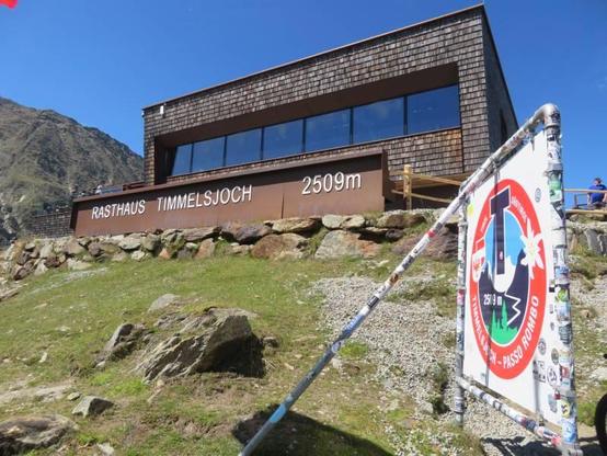 Aufnahme am Timmelsjoch, 18. August 2025: Rechts das dort aufgestellte Schild mit der Beschriftung "Tirol Südtirol Timmelsjoch - Passo Rombi" sowie "2509 m"; In der Mitte das Rasthaus mit der Beschriftung "Rasthaus Timmelsjoch 2509m"