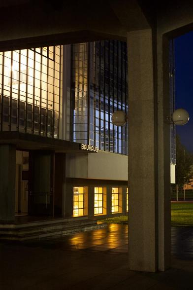 Light shines from inside through a glass facade. Each room has a different light color. The text “bauhaus” is written on the facade.