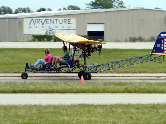 Two people sit it in a small, open-frame Breezy aircraft which has just landed at AirVenture 2008 in Oshkosh. The aircraft has a visible metal structure, a high wing, and a rear-mounted propeller engine. A traffic cone is nearby, and a hangar in the background displays the "AIRVENTURE OSHKOSH EAA" sign.