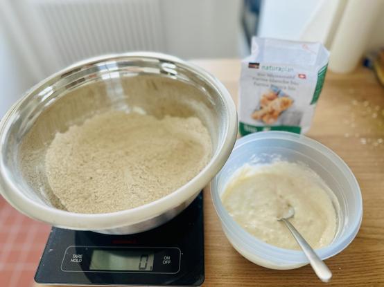 Ein grosse Aluschüssel mit gemischtem Mehl, daneben steht eine kleine Kunststoffschüssel mit einem Löffel drin. In der Schüssel blubbert der Sauerteig.