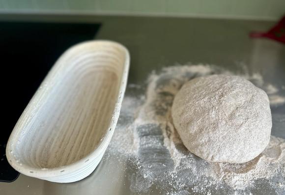 Eine Teigkugel liegt auf der bemehlten Anrichte, daneben steht ein längliches Gärkörbchen aus Bast.
