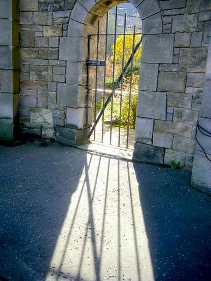 Strong sunlight shining through a garden gate, making a pattern on the pathway in front of it. Behind the gate you can see trees and plants.