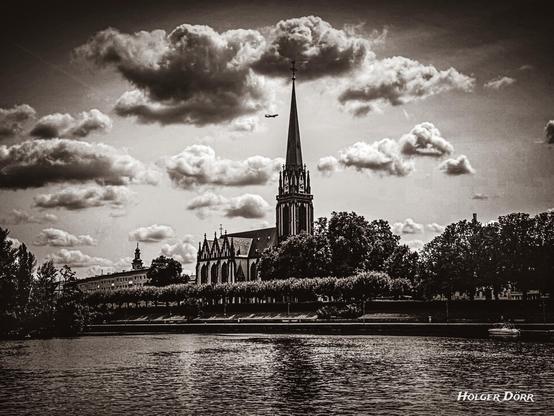 Schwarz-Weiß-Foto der Dreikönigskirche in Frankfurt am Main mit markantem Turm, Wolkenhimmel und Main im Vordergrund.