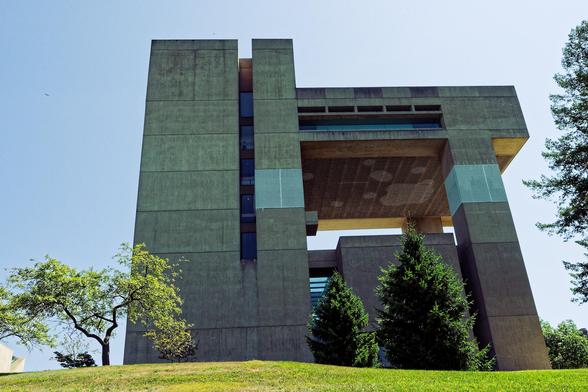 A large concrete building rises out of a close cropped grass lawn with a spreading deciduous tree and two evergeens in front of it and just a little bit of I think another evergreen coming in from the left.  The building itself has a convention tower to the left sidt with a vertical slot of windows towards the right side of that,  a canilevers structures extrnewds to the right supported by two pillars,  it has a big window and five thin slotted windows above it.