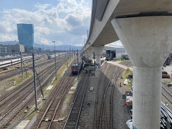 A field of tracks with a concrete viaduct and below a red and blue electric locomotive. 