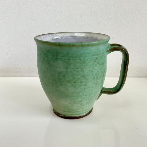 A shapely, handmade mug with greenish glaze on the outside, and white on the inside. It’s shown with an angled view against a white background.