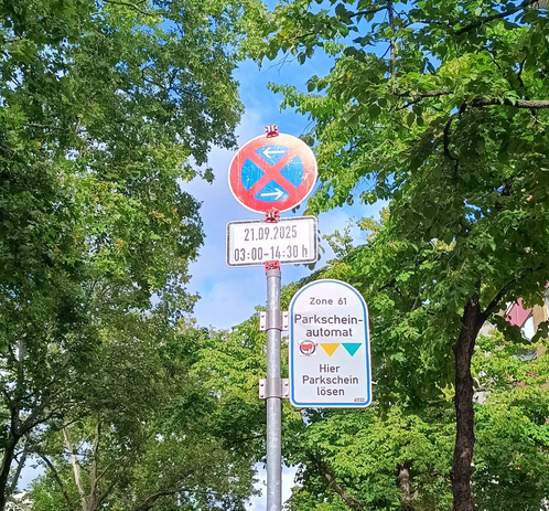 Das Bild zeigt eine Straßenanzeige in einer grünen Umgebung mit Bäumen im Hintergrund. Im Vordergrund befindet sich ein Metallpfosten mit mehreren Schildern. Das oberste Schild ist rund und rot mit einem blauen Kreuz und zeigt, dass Parken und Halten verboten sind. Darunter ist ein weißes Schild mit der Aufschrift "21.09.2025 03:00-14:30 h Zone 61", was die Gültigkeitszeit für die Parkregelung angibt. Ein weiteres Schild auf dem Pfosten weist auf einen "Parkscheinautomat" hin und gibt an, dass hier Parkscheine ausgegeben werden können. Die Schilder sind in deutscher Sprache und geben Informationen über die Parkregeln und die Verfügbarkeit eines Parkscheinautomaten an.