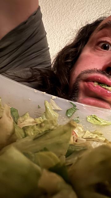 Das Bild zeigt einen Mann mit langen, dunklen Haaren und einem Bart, der in einem Selfie-Format aufgenommen wurde. Er hat einen überraschten oder amüsierten Gesichtsausdruck und blickt direkt in die Kamera. Seine Lippen sind leicht geöffnet, und es scheint, als würde er etwas von einem Salat essen, der in einem weißen Behälter vor ihm liegt. Der Salat besteht aus grünen Blättern, wahrscheinlich Blattsalat, und enthält auch andere Zutaten wie Tomaten. Der Hintergrund ist ein weißer, texturierter…