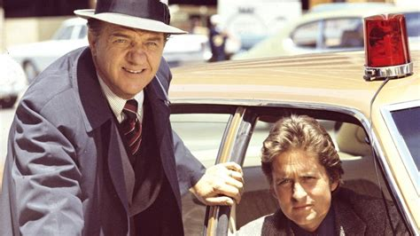 Snapshot from the TV series The Streets of San Francisco with Karl Malden standing outside of a yellow undercover squad car with a red bubble gum light on the roof, and with Michael Douglas sitting in the front passenger seat. Both gentlemen are looking towards the camera.