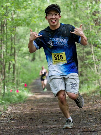 Asian man running with a black, blue and white shirt that says The SUNK CABBAGE and a yellow bib that reads 629 in black letters and grey shorts that say Homer Tennis.  One foot is in the ground and one is folded back and he's making hand signs with both hands.  He's running on a trail that is surrounded by thin little trees,  there are a few pink flags on the side of the trail and a woman in black clothers and seemingly bare shoulders is in the far distance.