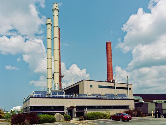 A low cream-colored building in front with a larger boxy building behind it,  two thin cream-colored stacks rise from that building while there are two (one obscured) red brick smokestacks behind it from the coal-fired system that it replaced.  Blue sky with cumulus clouds