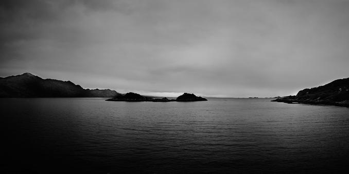 A monochrome image showing the calm waters of Loch Nan Uamh. The horizon is a blend of distant landforms and a cloudy sky. On the left, a dark hill slopes into the loch. In the foreground, rugged shorelines frame the water.