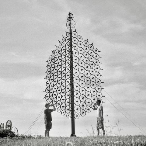 A black and white film photo. Two people have a drink in front of the giant L. J. Maasdam’s Wagon Wheel Sculpture in Grinnell, Iowa. Saturday, July 12, 2025.