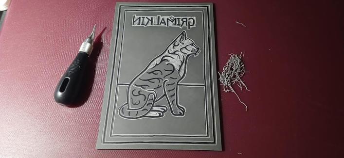 Image 1:

Picture of my "Grimalkin" cat being carved into lino. In the middle is the cat drawn onto lino with black permanent marker and shallow cuts being made around the black permanent marker. To the left is my cutting blade. To the right is a whole bunch of lino shavings shuffled into a pile for the photo.