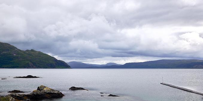 The image shows a view from Kilchoan harbour towards Mull. In the foreground, rocks and a small pier jut into the calm sea. Rolling hills stretch across the horizon under a vast, cloudy sky.