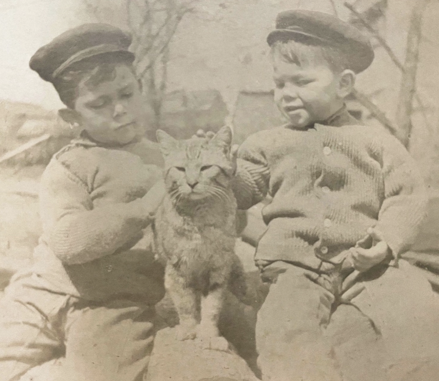Sepia toned black and white photo of two young white boys (possibly brothers based on resemblance) wearing cardigans and caps. Between them is a big happy, confident looking shorthaired tabby getting pets and attention from both boys.