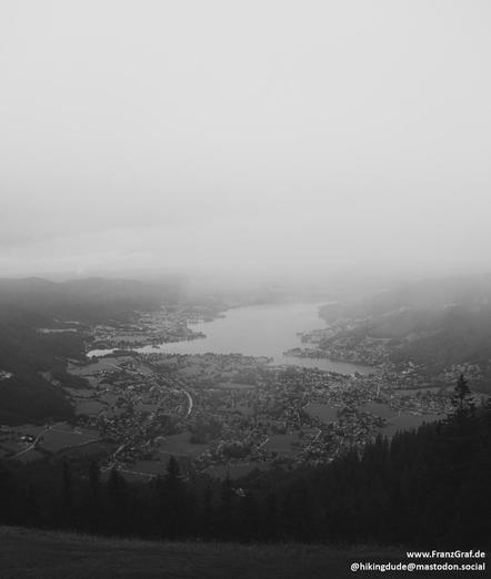 A serene black and white photo capturing the beauty of a lake nestled among a lush forest. The image showcases a tranquil scene with the still waters reflecting the surrounding trees. The misty atmosphere adds an ethereal quality to the landscape, enhancing the sense of mystery and tranquility. The dominant colors of grey and white create a soothing and timeless feel. This aerial view offers a peaceful escape into nature, inviting viewers to immerse themselves in the peacefulness of the scene. …