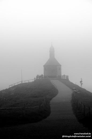 A black and white image depicting a building perched atop a hill, surrounded by fog and mist. A winding path leads up to the building, which is shrouded in a hazy atmosphere. The dominant colors in the image are white, black, and grey, creating a stark and moody landscape. In the foreground, a tower structure is visible, adding to the mysterious and eerie ambiance of the scene. The overall feeling of the image is one of solitude and isolation, with the building standing as a solitary beacon in …