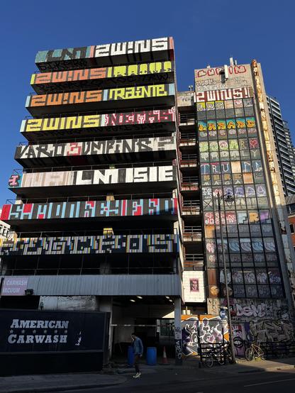 Close up shot of graffitied multistorey carpark, taken at ground level. It fills almost the whole picture, with only a little bit of a skyscraper and the dark blue spring afternoon sky in the background.