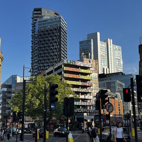 Various road traffic lights in the foreground, followed by a spring-green tree, then right in the centre of the image a narrow, graffitied multistorey carpark, with various glass London skyscrapers in the background.