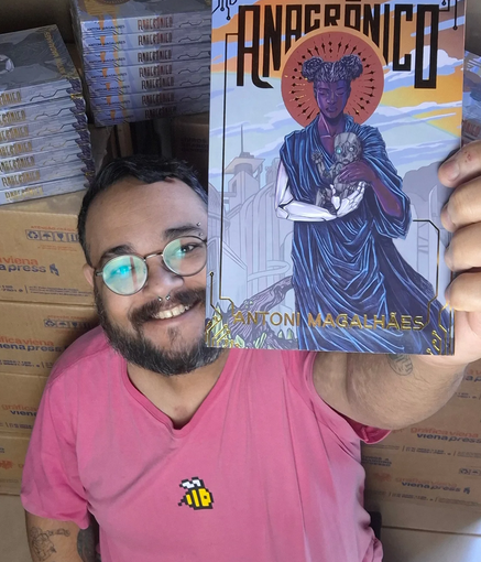 Foto do autor Antoni Magalhães sorrindo, segurando o livro Anacrônico perto da câmera. Ele é gordo, de pele branca, cabelos curtos preto e barba e bigode. Usa óculos redondos, tem um piercing no nariz, e veste uma camisa rosa com apenas o desenho de uma abelha amarela no peito. Atrás dele, várias caixas com exemplares do livro.