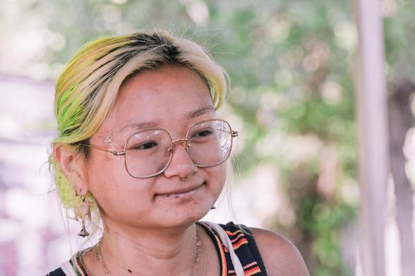 A woman with short-to-medium-hair combed back with yellow and green color and large squarish-round glasses has a gaint smile,  behind her spots of lights can be see shining through trees