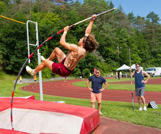 A man wearing a tan tank top and maroon shorts and white shotes and a headband has hair in tight ringlets and is holding on to a pole bent almost horizontal at the top as he ascends towards the yellow bar partially visible on the upper left,  below him is a big red and white mat.  A coach looks on wearing dark glasse, a blue shirt and pale red shorts with his hands on his hips together with a man holding on to  short pole,  behind them is the curve section of a red track with a few runners stan…