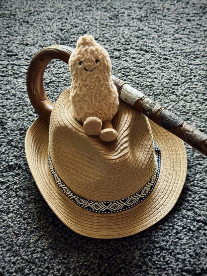 Auf dem Boden ein Strohhut, auf dem eine Plüscherdnuss sitzt. Ein Wanderstock liegt schräg auf dem Hut.