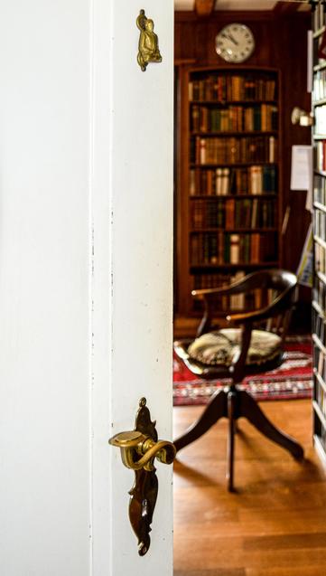 Ein leicht geöffneter weisser Türrahmen im Vordergrund, der eine altmodische Messing-Türklinke und einen Türklopfer zeigt. Dahinter ist ein Raum zu sehen, der wie eine Bibliothek oder ein Arbeitszimmer aussieht, mit Bücherregalen und einem hölzernen Schreibtischstuhl. Im Hintergrund hängt eine Wanduhr.
