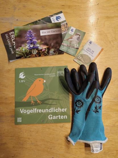 Das Schild "Vogelfreundlicher Garten"  vom LBV und dem Bayrischen Landesamt für Umwelt, zusammen mit Broschüren zu Gartenvögeln und Igeln und einem Paar Gartenhandschuhe.
Der Makro-Hortus wurde heute als vogelfreundlicher Garten ausgezeichnet.