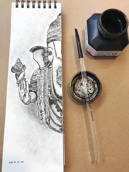 Black ink brushwork representing half of Ganesh on a talk and narrow drawing pad. The ink bottle and flat brush are at its side