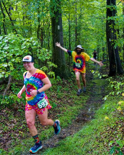 Three runners on a trail between two trees with square white blazes.  In front a woman with a billed cap, big smile, tie dyed shirt and crazy shorts and socks with a green bib that says 593 and behind her a man with a beard and boonie cap also with a tie dyed shirt and his arms spread out diagonally and a third runner much further away who seems more subdued.