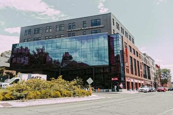 A 7 story or so broad building had brown bricks at the the top and a sign that reads THE STUDENT AGENCY BUILDING,  below that the front has a glass facade that reflects a sky with clouds and towards your right,  a red brick facade.  In front find a Hertz rental van,  a garden of brownish green (color graded) foliage and a road.  Above:  a subdued (color graded) blue sky with a few clouds,  Behind: other buildings a little less tall and some parked cars.