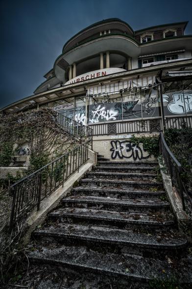 Verwilderte Treppenstufen führen zu einem verlassenen, mehrstöckigen Gebäude, dessen Fassade mit Graffiti  bedeckt ist. Die Schriftzüge "HIRSCHEN" und "RESTAURANT" sind zu erkennen. Efeuranken ranken sich an den Wänden empor, während die metallenen Geländer der Treppe von Rost gezeichnet sind. 
