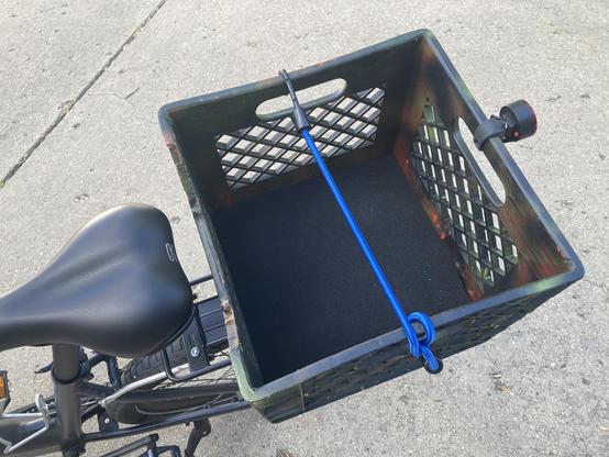 A milk crate on a rear bike rack. There is a piece of foam at the bottom of the crate and a bungee cord attached. 