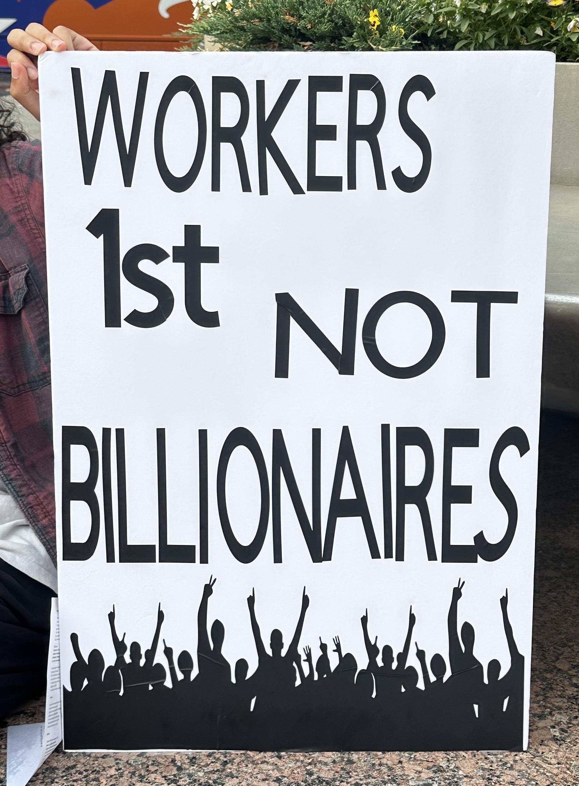 A person holding a protest sign that reads "WORKERS 1st NOT BILLIONAIRES" with silhouettes of raised fists along the bottom.