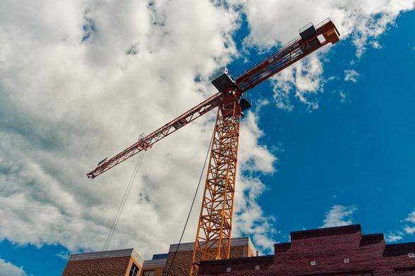 A yellow crane rises vertically straight up with a space frame beam supporting a cross beam long in towards the left and with a counterweight towards the right,  above a blue sky mostly obscured by a huge cumulus cloud,  in the bottom right the very tops of a few buildings are in front of or behind the crane.