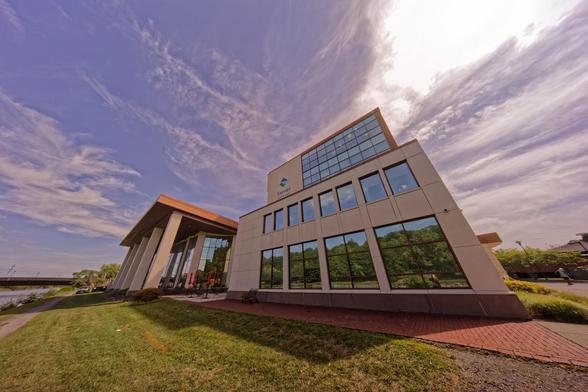 A large building looks small and strangely distorted:  to the left a smaller box is stacked on a larger box,  both with a creamy colored wall broken by windows that reflect the sky,  to the left an all-glass section is covered by an awning with vast pillars -- in front close cropped grass,  a brick path and a bridge,  above a blue sky with clouds illuminated by the sun at the upper left,  making the building cast strong shadows to the lower right.
