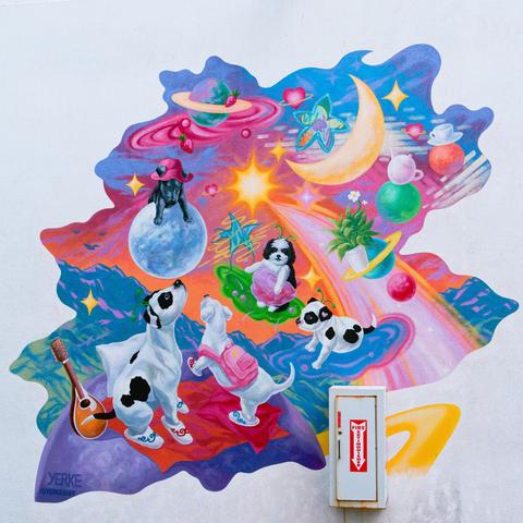 A mural on a wall only slightly marred by a white box for a fire extinguisher with a red sticker with a white arrow that points down.  The mural is irregularly shaped and has patches of blue water and purple sky with moons and planets and shooting stars and lots of white dogs with red spots not to mention whimsical things like tea cups and stringed instruments