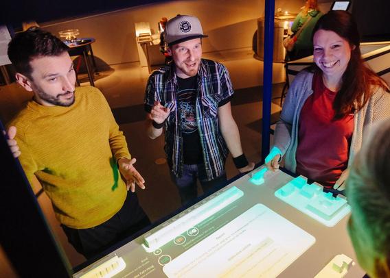 Drei junge Erwachsene stehen gemeinsam an einem interaktiven Tisch mit leuchtenden Bedienelementen. Sie diskutieren und lachen, während sie mit digitalen Objekten und Projektionen auf der Oberfläche experimentieren.