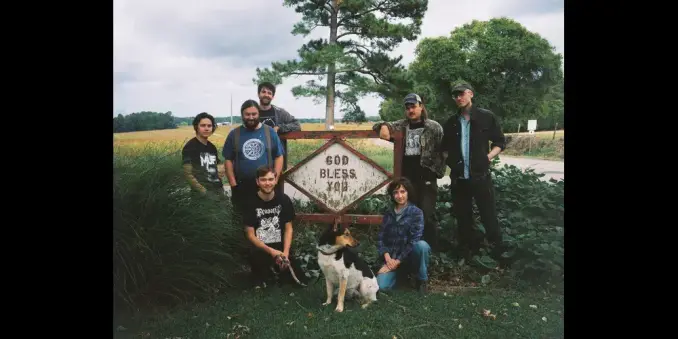 The Weirs collective and a dog pose by a "God Bless You" sign in a rural setting. They're dressed casually, with trees and fields in the background.
