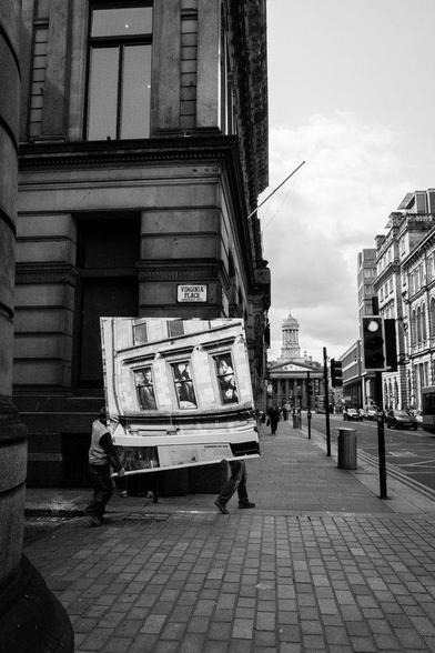 Two workers moving a very large mirrored surface by hand on a street in central Glasgow. In the mirror you can see windows from the building across the street, which is not in the photo. Black and white.