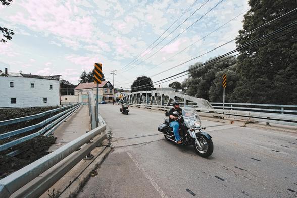 We're looking almost straight down a small bridge with metal struts, sidewalks and double guardwails on both sides,  two motorcyclists are driving towards us,  one just got off the bridge and the other is in the middle of it;  there are many electric wires running over and parallel to the bridge and on the other side of the creek is a white building with four windows is on the left  and straight down the bridge a yellow building,  to the right some large deciduous trees