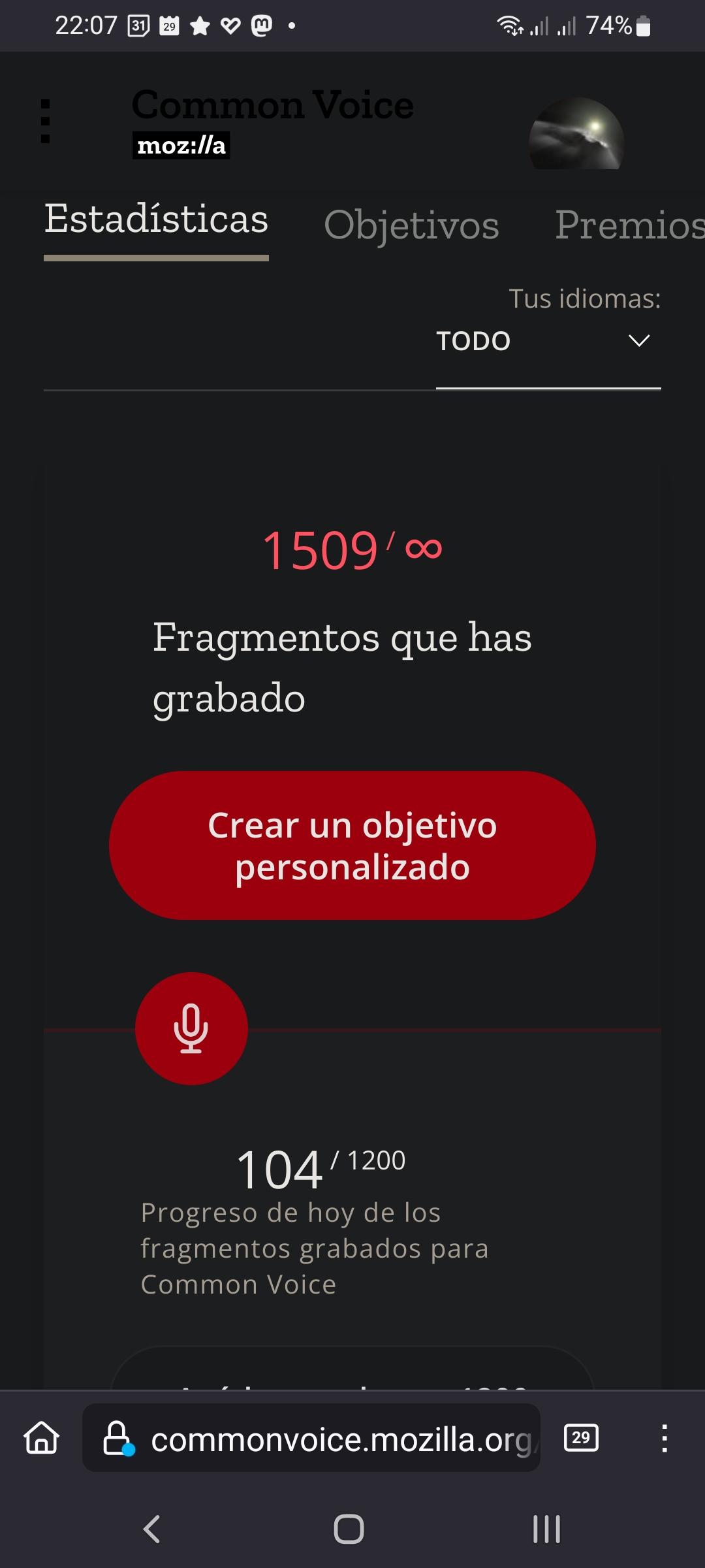Screencap shows a mobile device screen displaying the Common Voice website. The top of the screen shows the time as 22:07 and the battery level at 74%. The website's title "Common Voice" is prominently displayed, with a navigation menu on the left and a user profile icon on the right. The main section of the screen is titled "Estadísticas" (Statistics), with options for "Objetivos" (Objectives) and "Premios" (Awards) below it. The user's languages are set to "TODO" (All). The screen highlights that the user has recorded 1509 fragments, with a red button labeled "Crear un objetivo personalizado" (Create a personalized goal). Below, a progress bar shows the user has completed 104 out of 1200 fragments for the day. The website's URL, "commonvoice.mozilla.org," is visible at the bottom of the screen.