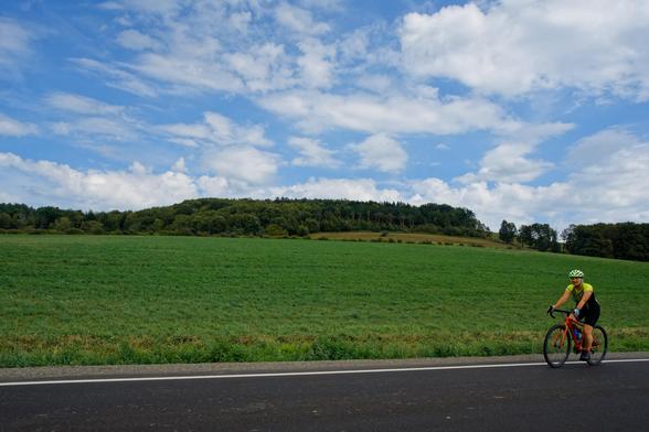 A woman rides a bicycle on the far right of the frame towards the left on a road that takes up maybe 1/10 of the frame,  the next 1/3 is a green grass field,  and above that a forested hill with another grass field cut out of it,  and above that a sky with white cumulus and cirrus clouds
